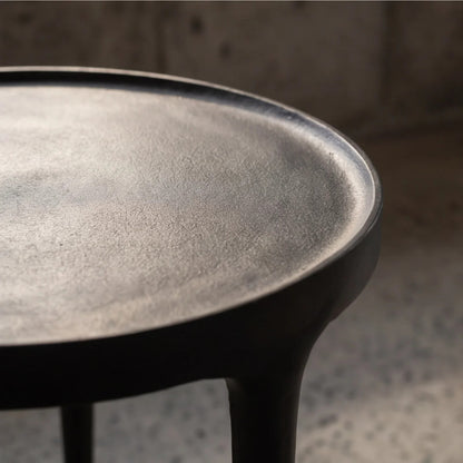 Close-up of a black side table top with a textured matte finish and a thick rim, supported by dark legs, set against a blurred concrete background.
