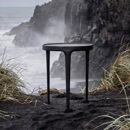 A round black side table with three tapered legs stands on a black sand beach with sea grass and a dramatic rocky coastline in the background.