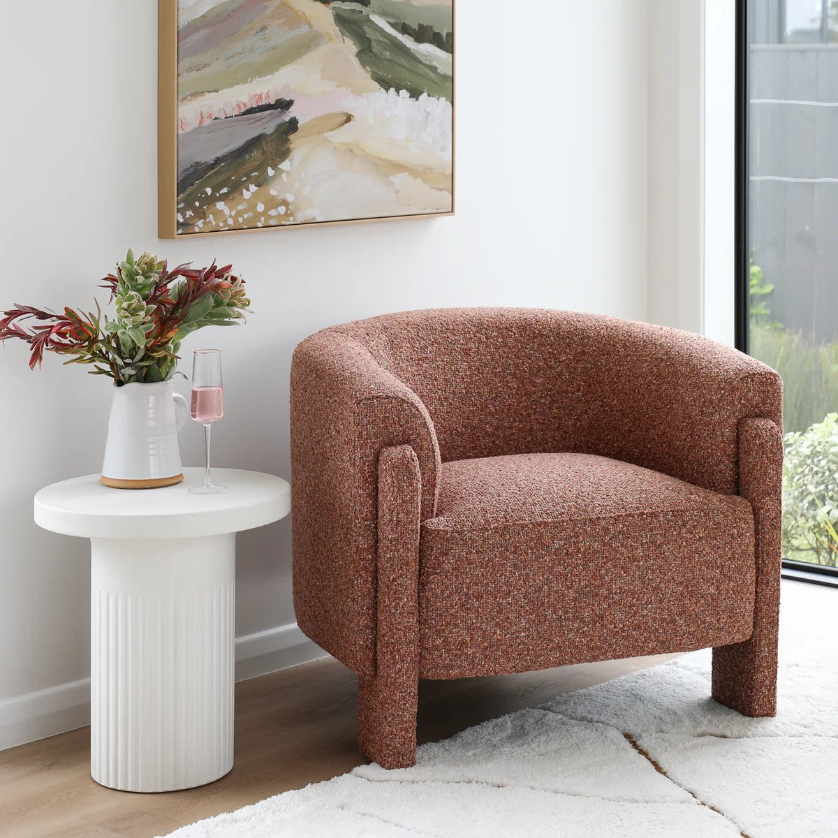 Blush armchair in a room with a white side table, flowers, and a painting on the wall.