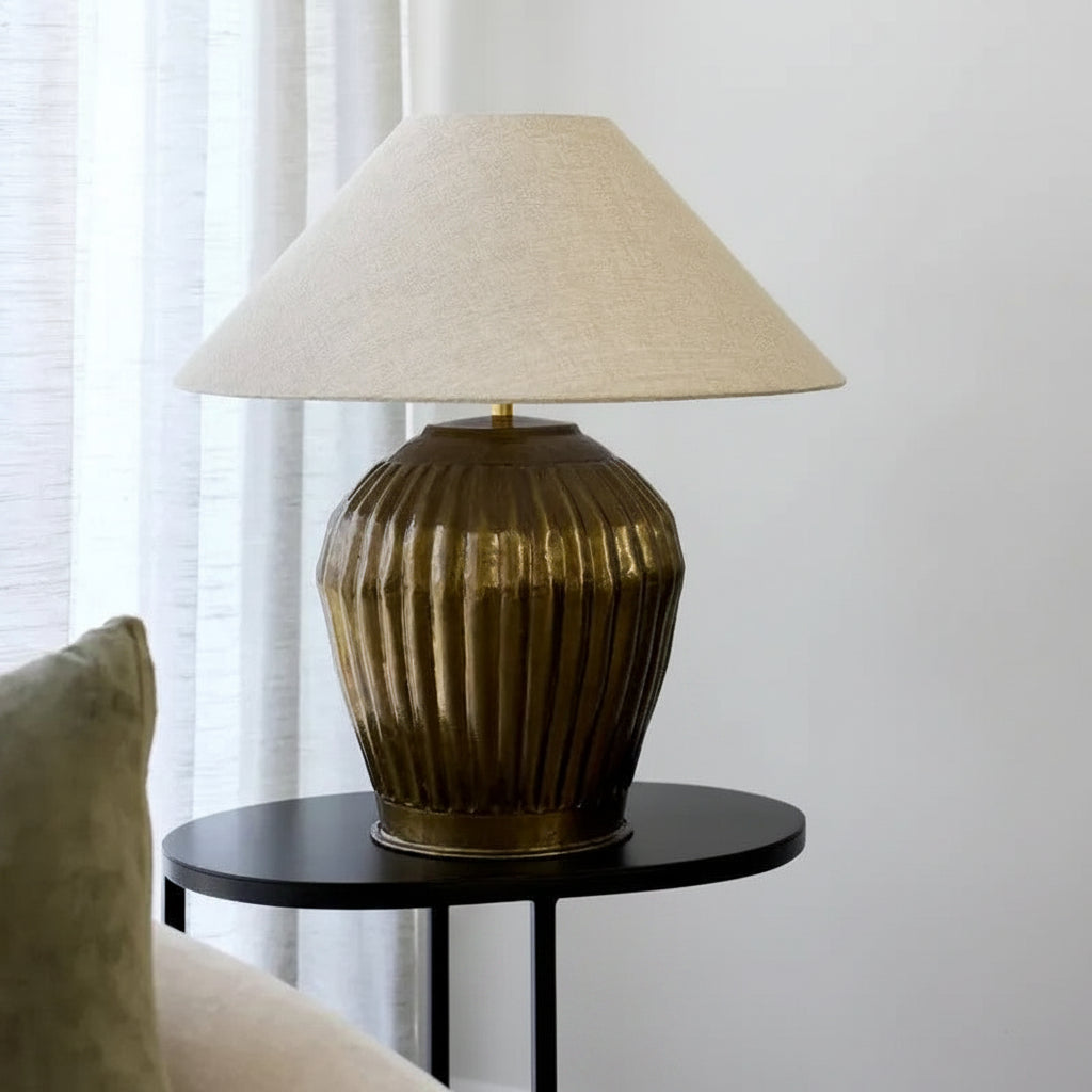 Antique brass table lamp with a linen shade on a black side table against a white curtain 