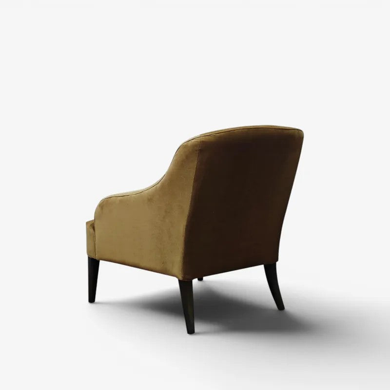 A single upholstered armchair shown from the back on a side angle, in a studio setting, featuring warm gold/tan velvet upholstery and dark tapered legs on a white background.