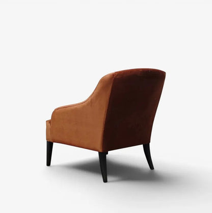 A single upholstered accent chair shown from the rear three-quarter angle on a clean white studio background. The chair has a warm rust-brown velvet seat and back, with slim dark legs.