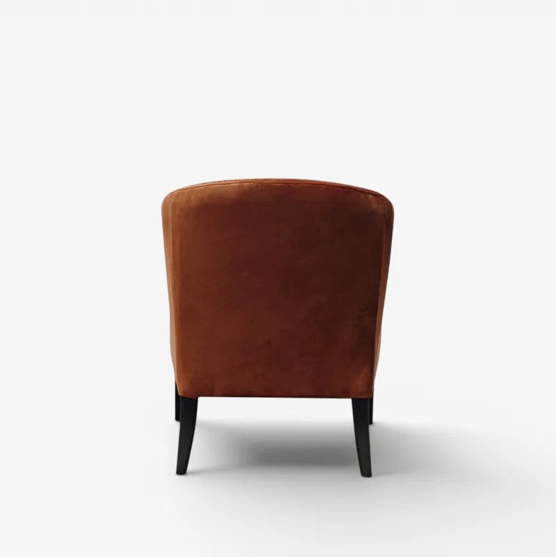 A single upholstered armchair shown from the back, featuring a warm rust-brown velvet seat and back cushion with slim black legs, set against a clean white studio background.