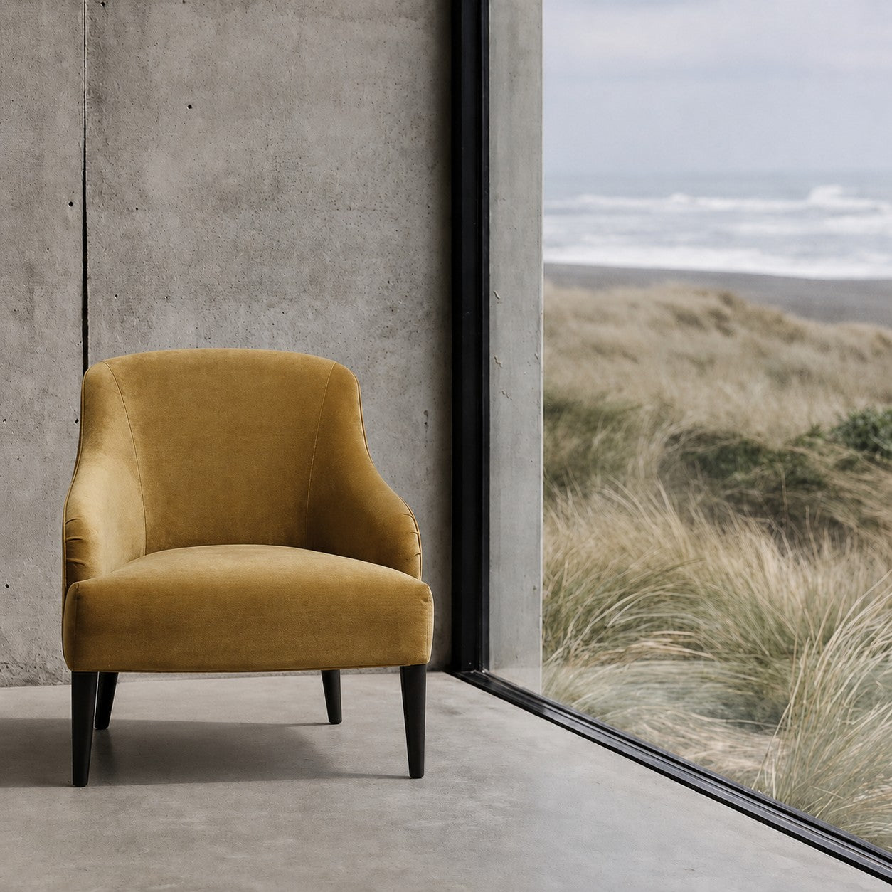 A gold/tan velvet armchair with dark legs sits in a minimalist room with a concrete wall and a large floor-to-ceiling window overlooking a beach dune landscape.