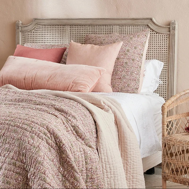 A styled bed with a distressed light grey wooden cane headboard, layered with dusty pink and blush velvet pillows, a textured pink quilt, and white bedding, set against a warm beige wall.