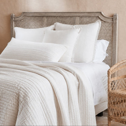 A styled bed with white textured bedding, including a quilted white duvet and matching pillows, set against a rustic grey washed headboard with woven cane panels. A matching woven chair is partially visible on the right against a textured beige wall.