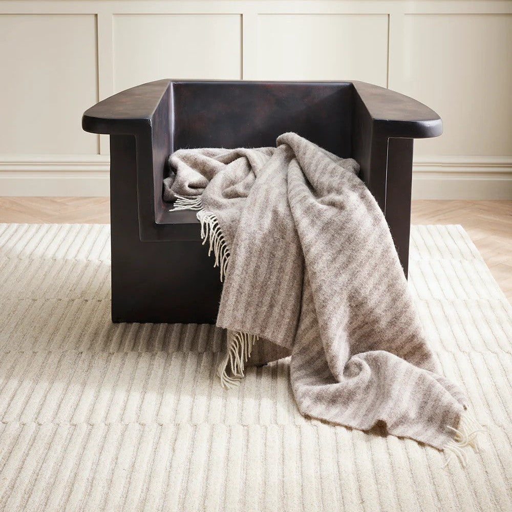 Dark wooden armchair with a oatmeal  throw blanket on a light coloured rug against a neutral wall.