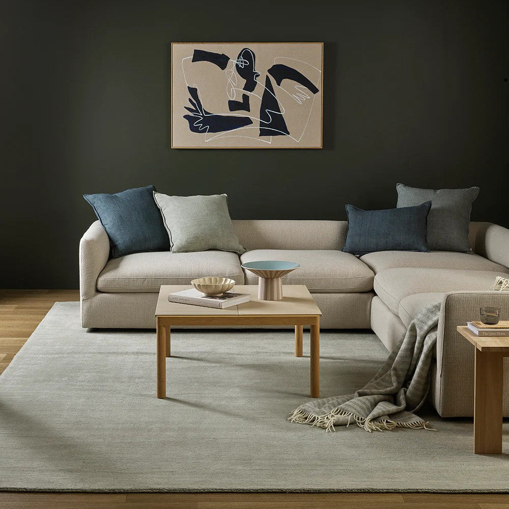 Modern living room with a beige sectional sofa, wooden coffee table, abstract art on the wall and a silver grey throw blanket draped over the sofa.