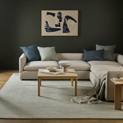 Modern living room with a beige sectional sofa, wooden coffee table, abstract art on the wall and a silver grey throw blanket draped over the sofa.