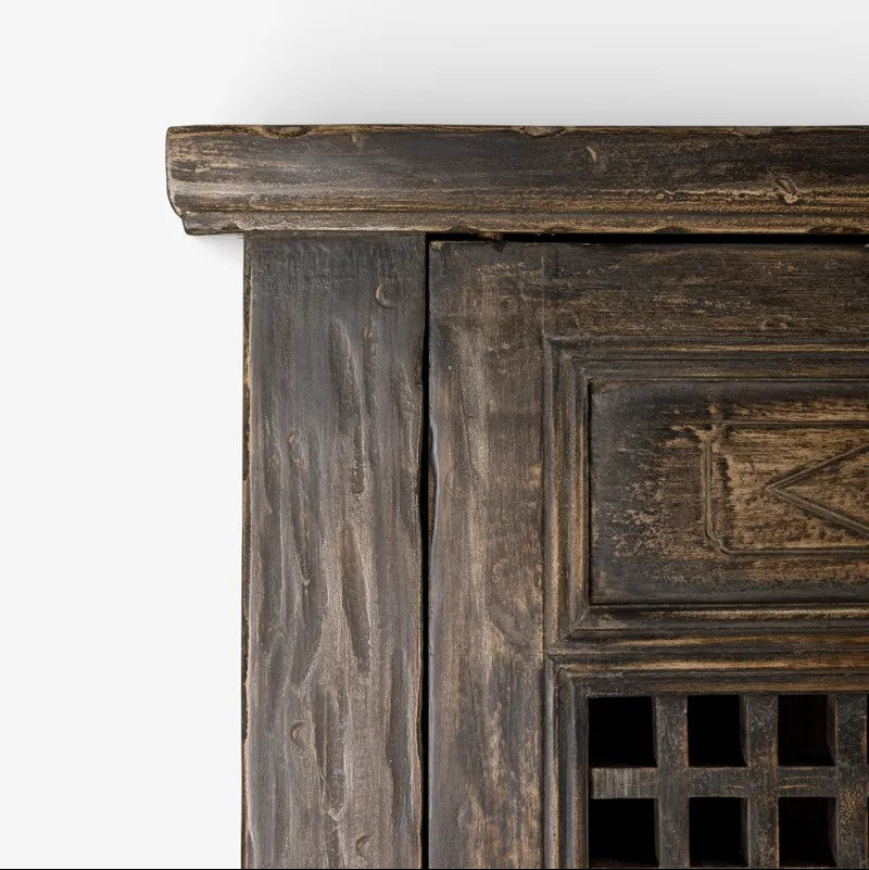 Close-up of a dark rustic wooden cabinet corner with carved panel detailing and a lattice window, set against a plain white background.