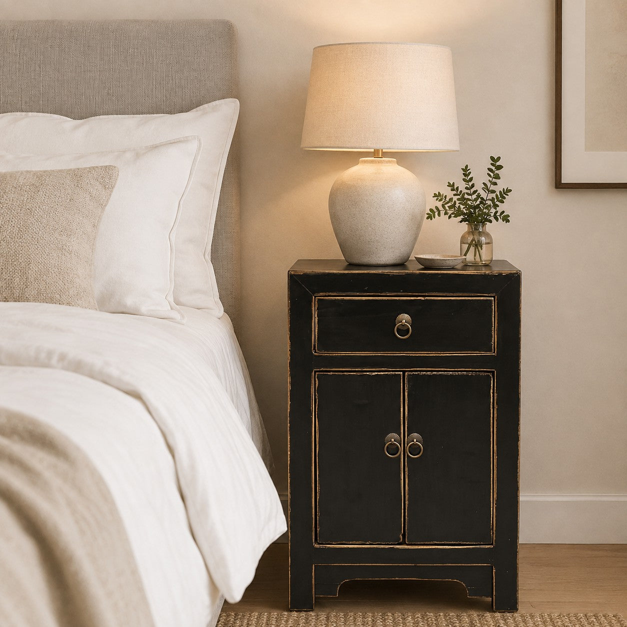 A black bedside cabinet with natural edges and ring pulls stands beside a bed with white linens in a softly lit bedroom. A beige table lamp and a small vase with greenery sit on top.
