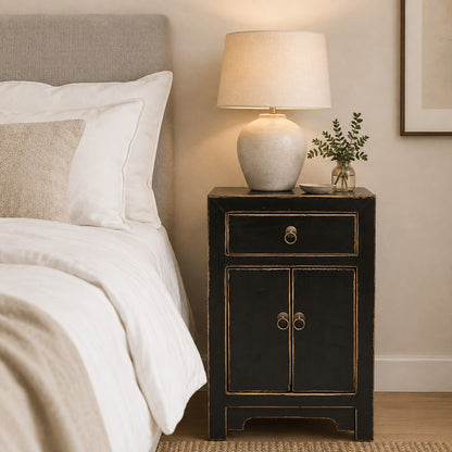 A black bedside cabinet with natural edges and ring pulls stands beside a bed with white linens in a softly lit bedroom. A beige table lamp and a small vase with greenery sit on top.