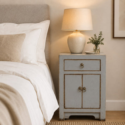 A grey painted bedside cabinet in a bedroom setting, styled with a lit table lamp and a small vase of greenery and bedding