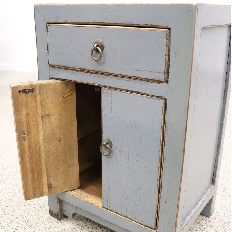 A small distressed grey wooden bedside cabinet with a single drawer and a lower door, shown with the door open to reveal the interior. The cabinet has ring pulls and is photographed against a white wall and speckled concrete floor.