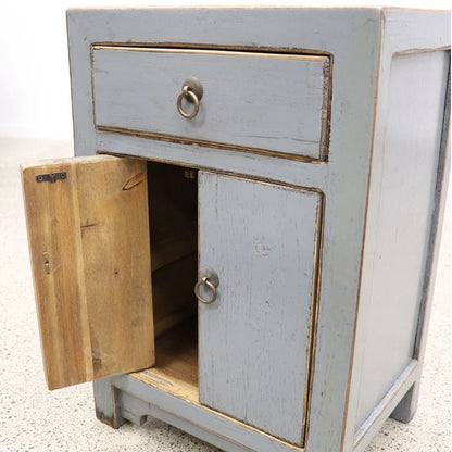 A small distressed grey wooden bedside cabinet with a single drawer and a lower door, shown with the door open to reveal the interior. The cabinet has ring pulls and is photographed against a white wall and speckled concrete floor.