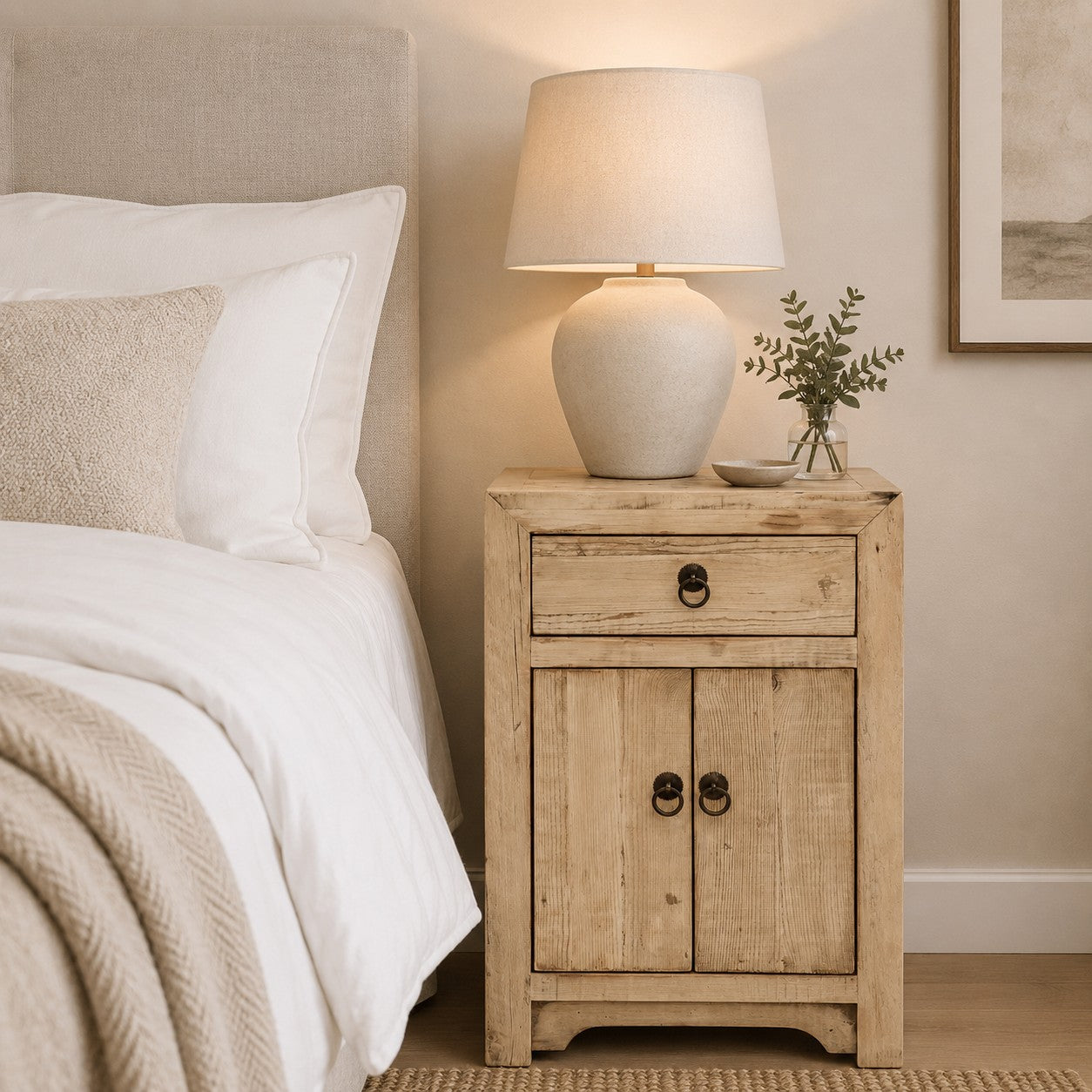 A rustic light wood bedside cabinet  in a bedroom setting, styled with a ceramic table lamp, a small vase of greenery, and a bed with white linens beside it.