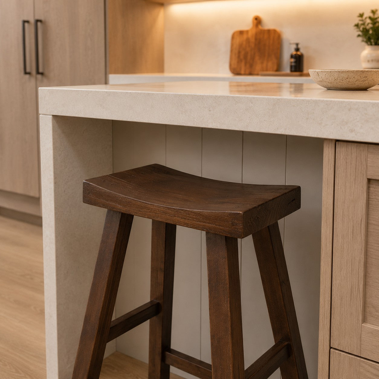 A dark brown wooden bar stool with a curved seat, positioned at a kitchen island with a light beige stone countertop and light wood cabinetry.