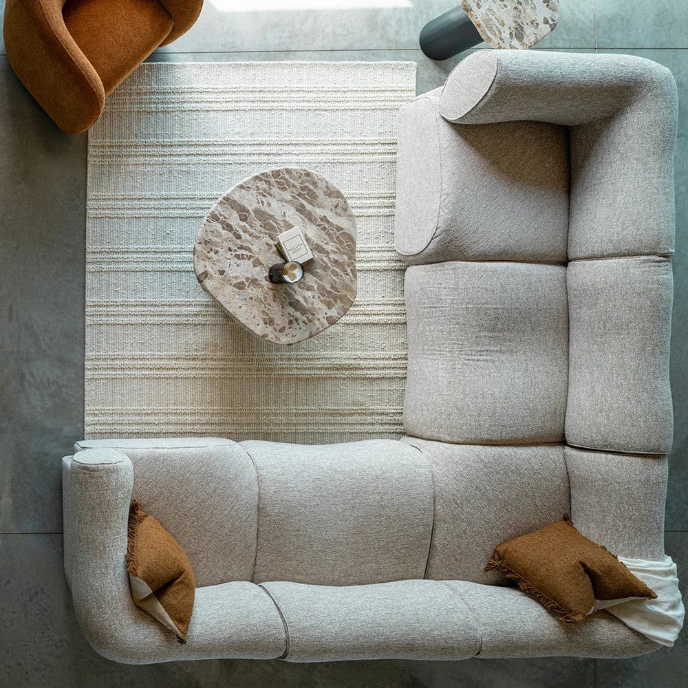 An overhead view of a modern modular sectional sofa in a light beige textured fabric, styled with two brown accent pillows on a striped rug with an organic marble top coffee table.