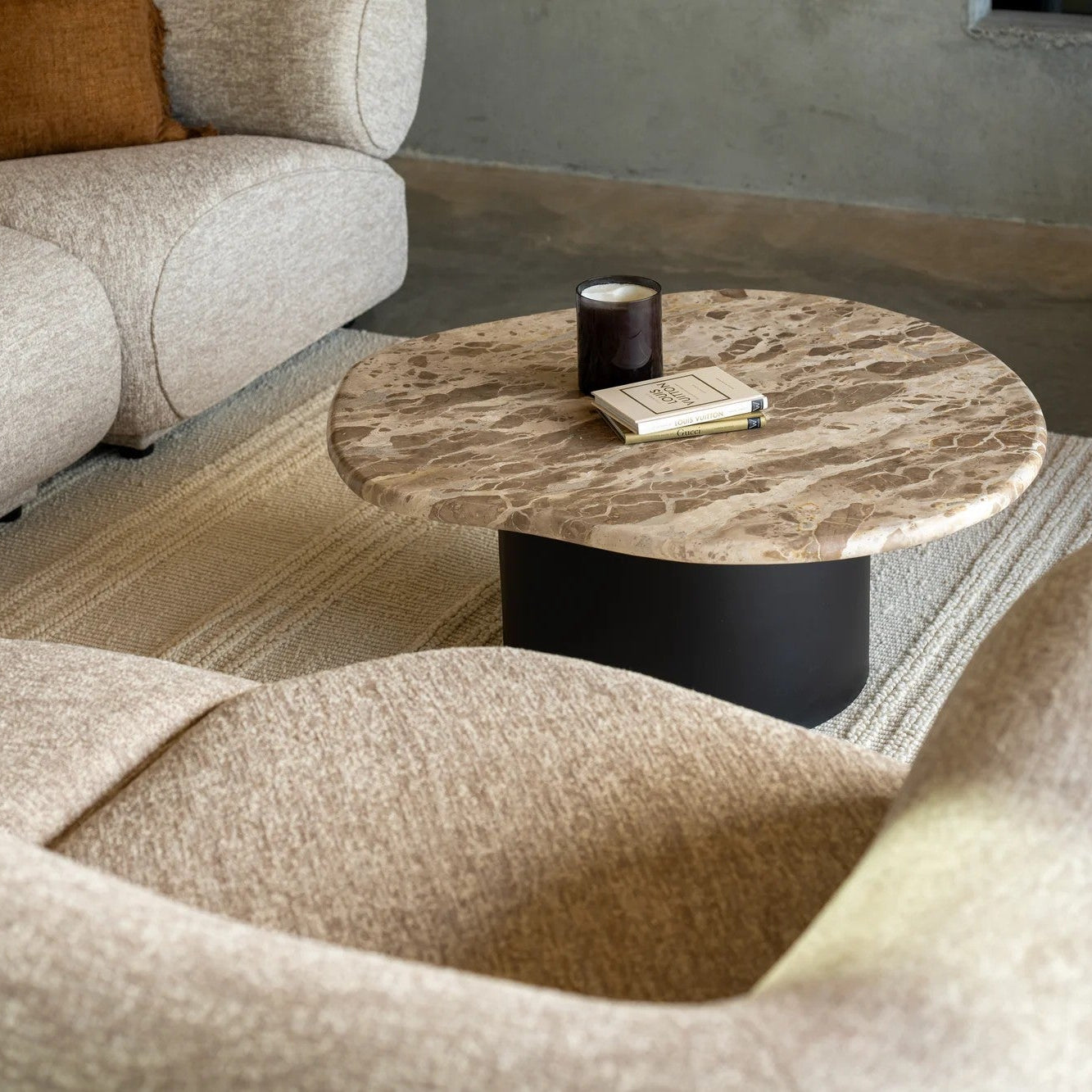 A modern living room scene featuring a curved, low-profile beige upholstered Corner suite sofa, and a organic marble top coffee table with a black pedestal base.