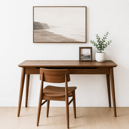 A mid-century modern wooden writing desk paired with a matching wooden chair, styled in a minimalist room with a framed beach print on the wall, a small picture frame, and a potted plant.