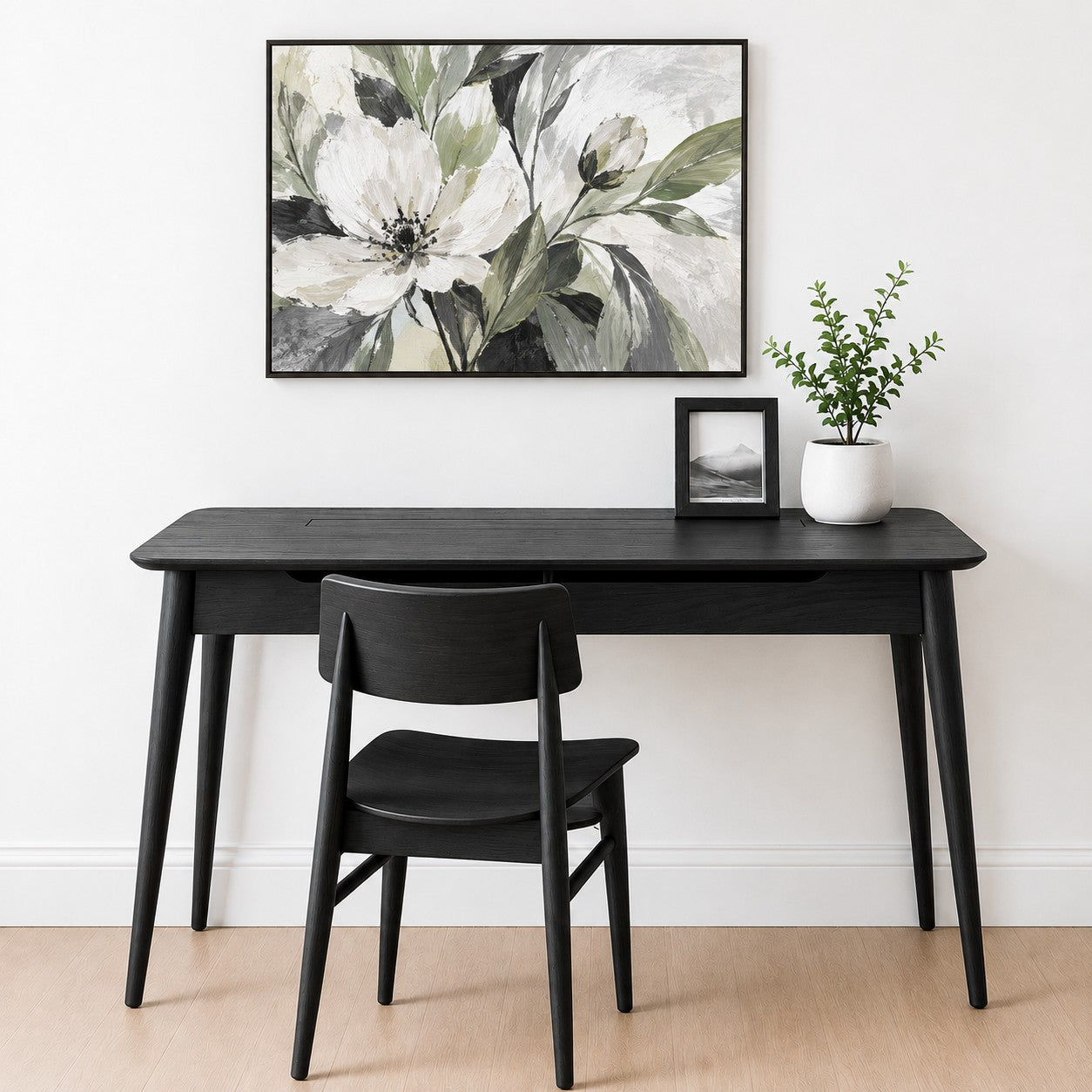 A black wooden writing desk with matching black chair, styled in a minimalist room with a white wall, a framed floral print, a small potted plant, and a picture frame.