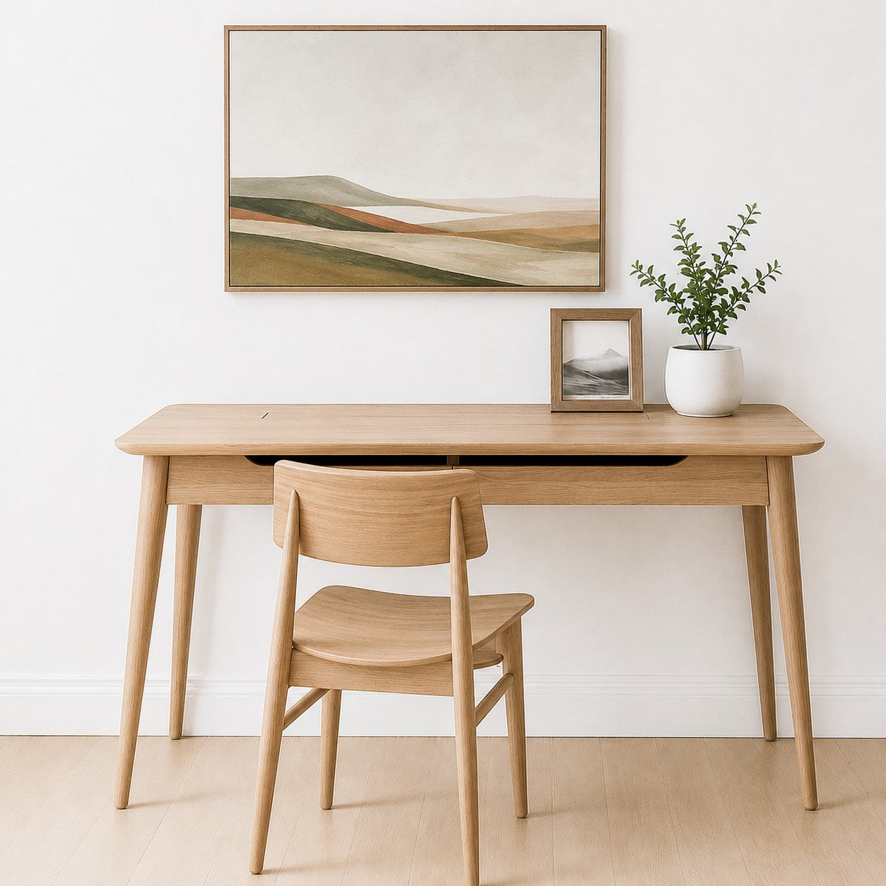 A light natural wood writing desk with tapered legs and a matching wooden chair, styled in a minimalist room with a white wall, a framed landscape painting, and a small potted plant.
