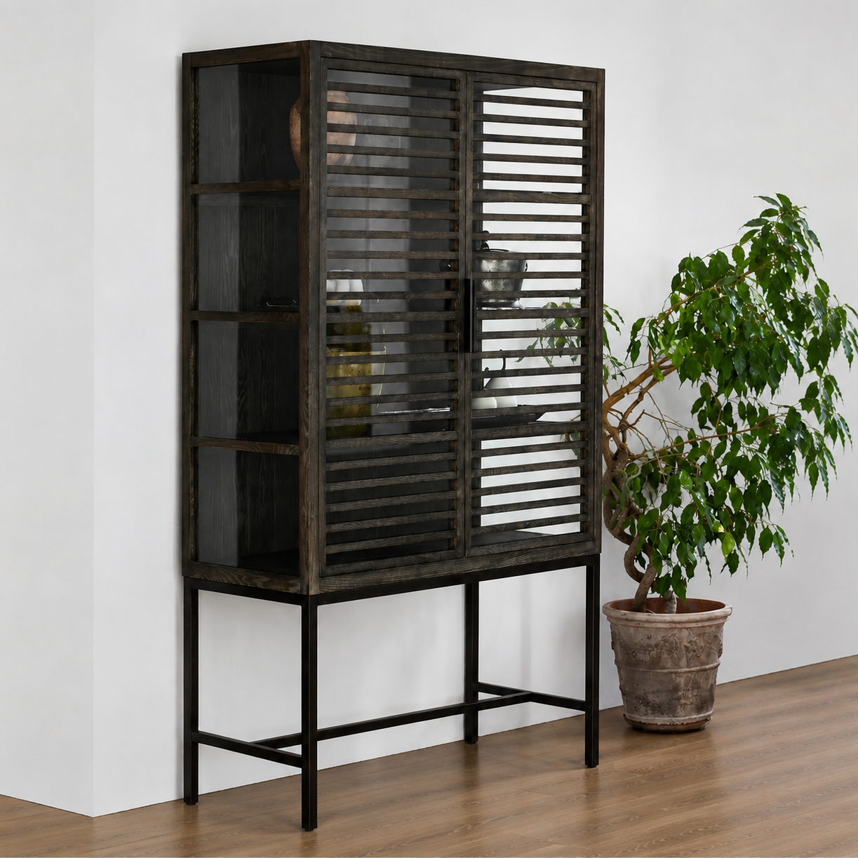 A tall dark wood cabinet with slatted doors and a black metal base, shown in a room setting next to a potted green plant on a wooden floor.
