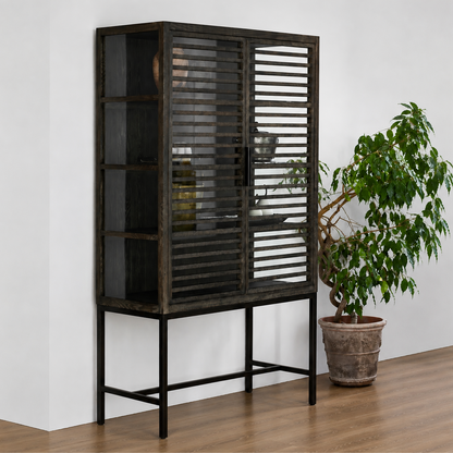 A tall dark wood cabinet with slatted doors and a black metal base, shown in a room setting next to a potted green plant on a wooden floor.