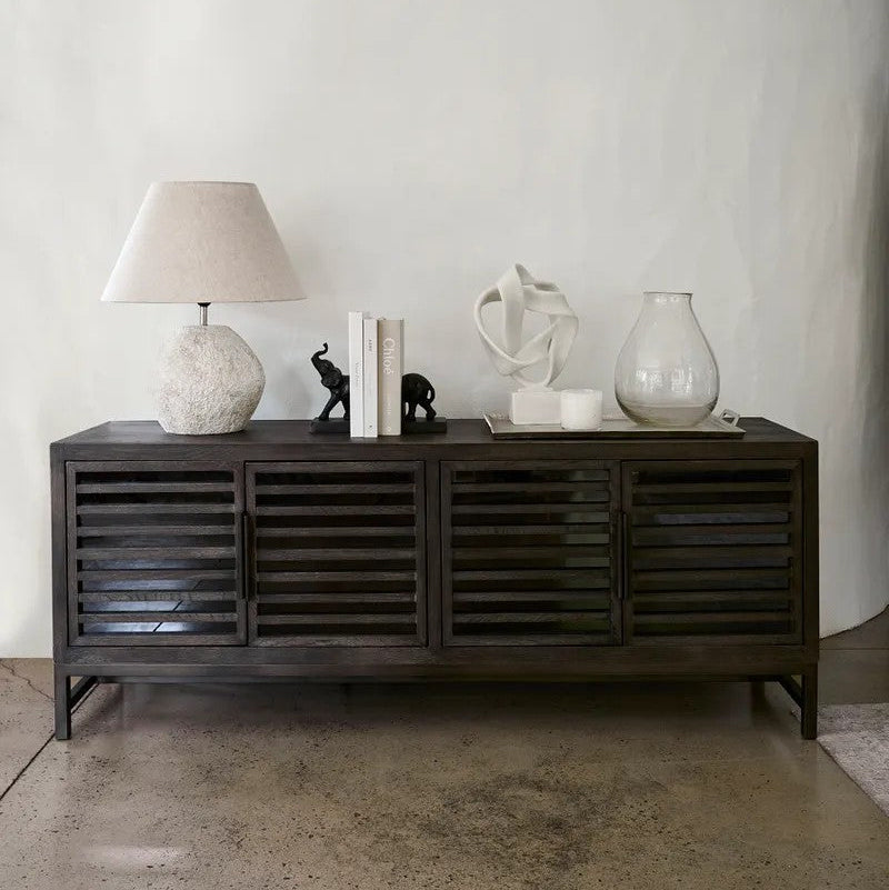 A dark grey wooden sideboard with slatted doors, styled with a textured white lamp, books, small elephant figurines, a white sculptural object, and a glass vase against a white wall and concrete floor.