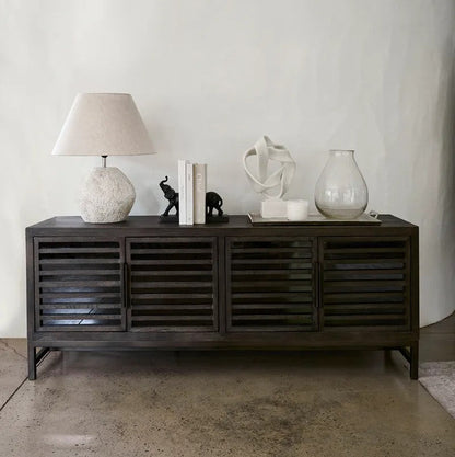 A dark grey wooden sideboard with slatted doors, styled with a textured white lamp, books, small elephant figurines, a white sculptural object, and a glass vase against a white wall and concrete floor.