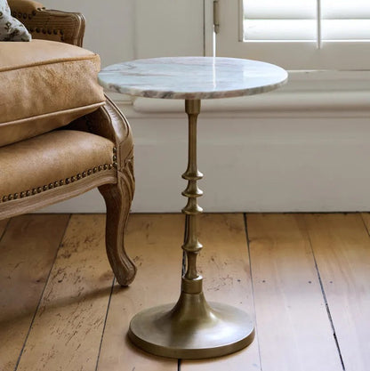Small round marble-top table with brass base next to a beige armchair on wooden floor.