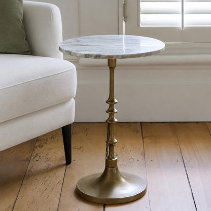 A round marble-top side table with a flared brass pedestal base, shown in a bright living room next to a white armchair on a wooden floor.