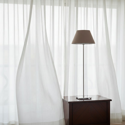 Table lamp with a brown shade sitting on dark bedside cabinet with white billowing drapes 