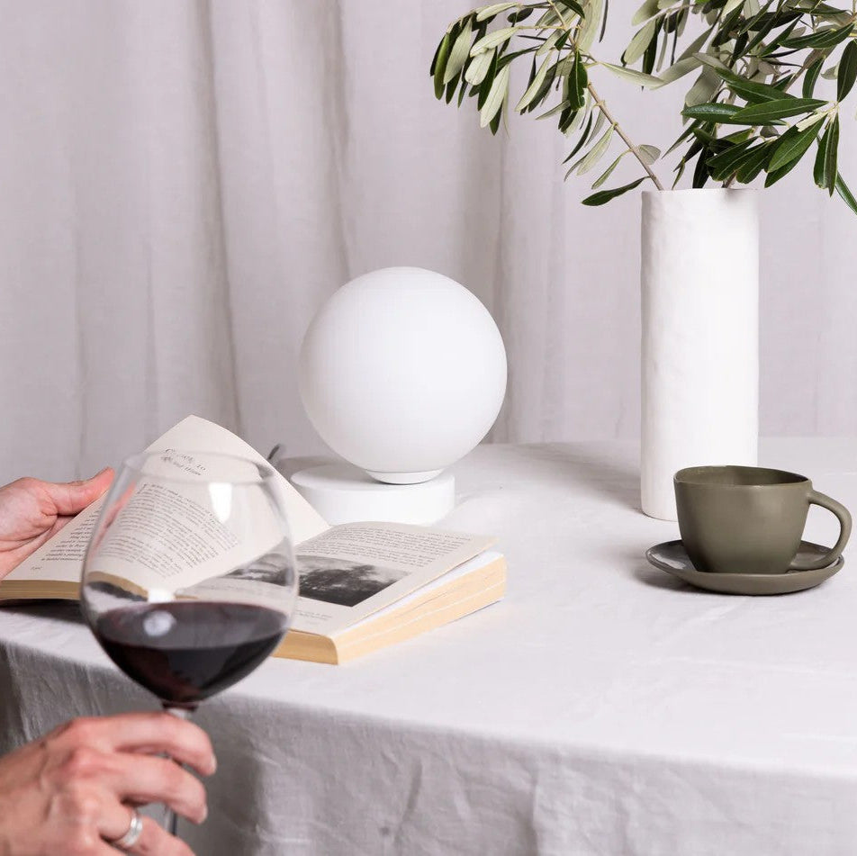 Person holding a glass of red wine at a table with a book, cup, and decorative items.
