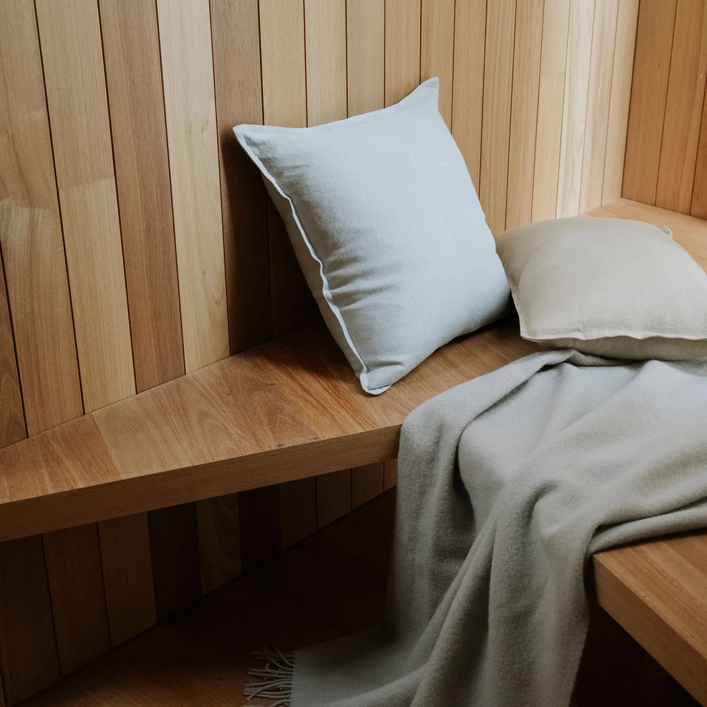 Wooden bench with light grey cushions and a blanket against a wooden wall.