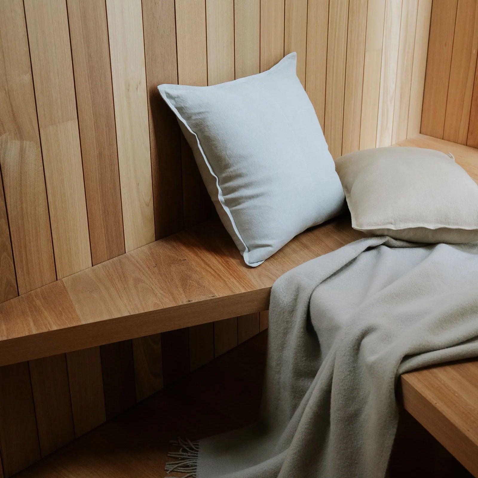 Wooden bench with light grey cushions and a blanket against a wooden wall.