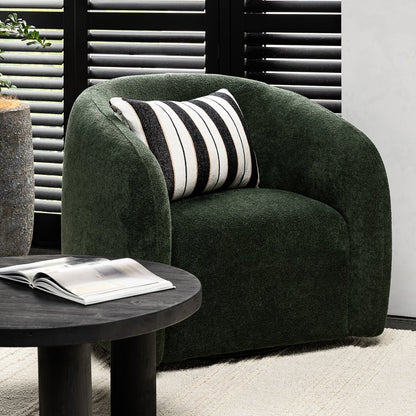 Green armchair with a striped cushion next to a round dark wood side table in a room with white walls and black shutters.